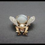 Broche oro amarillo y blanco 18k con forma de insecto decorado con piedra gris y perla blanca, las alas van cuajadas de puntas de brillantes